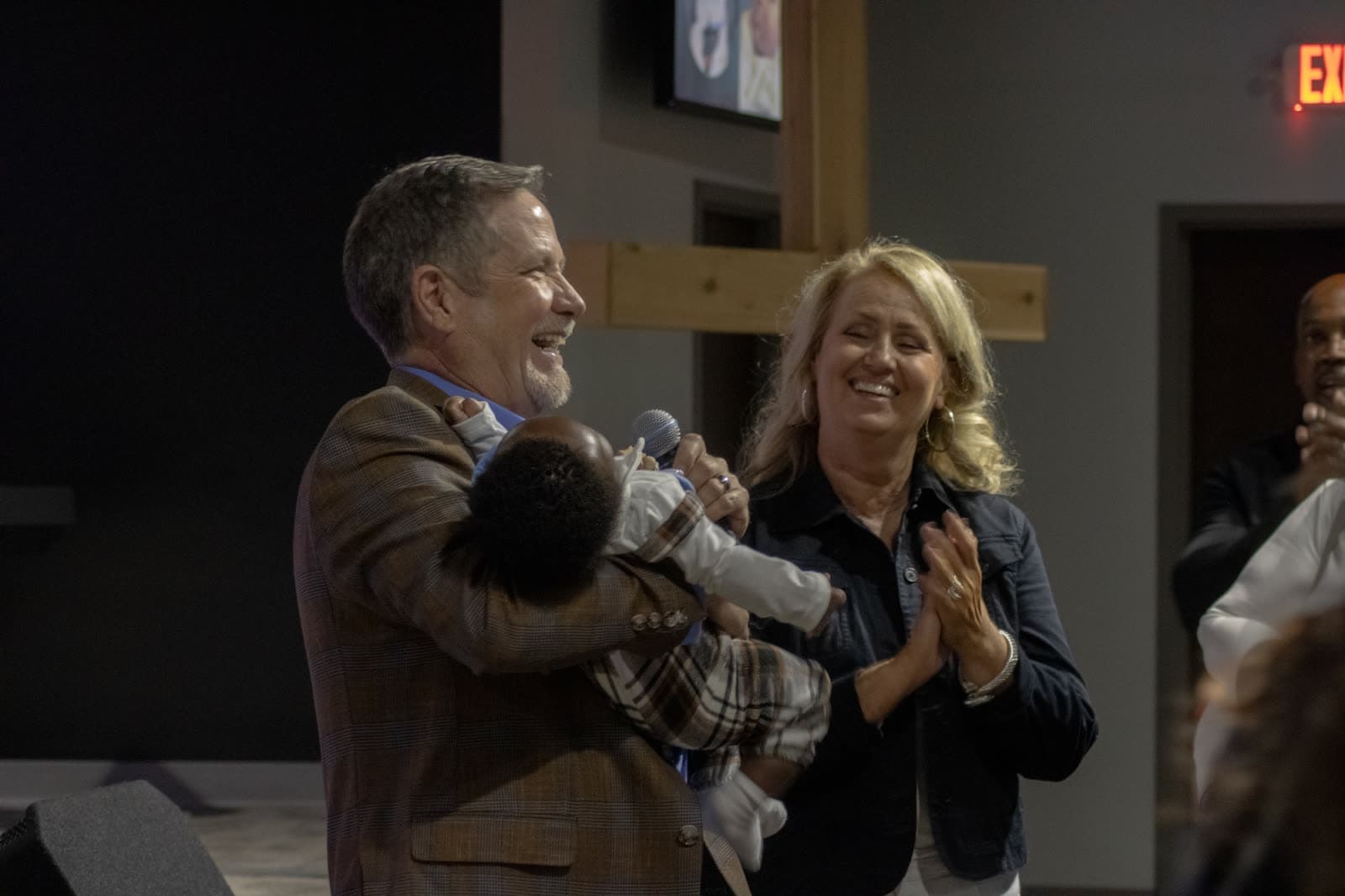 A pastor laughing as he holds a baby during a dedication at CrossOver, with a woman beside him clapping